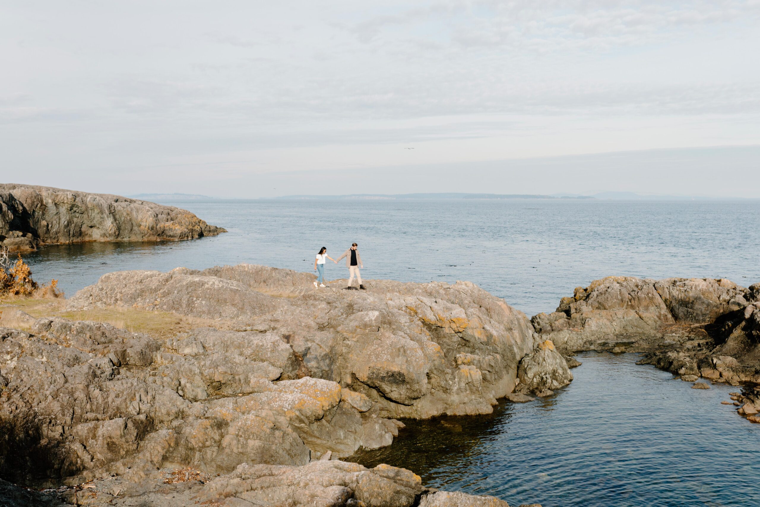 A man and woman are holding hands and standing on rocks overlooking an endless body of water