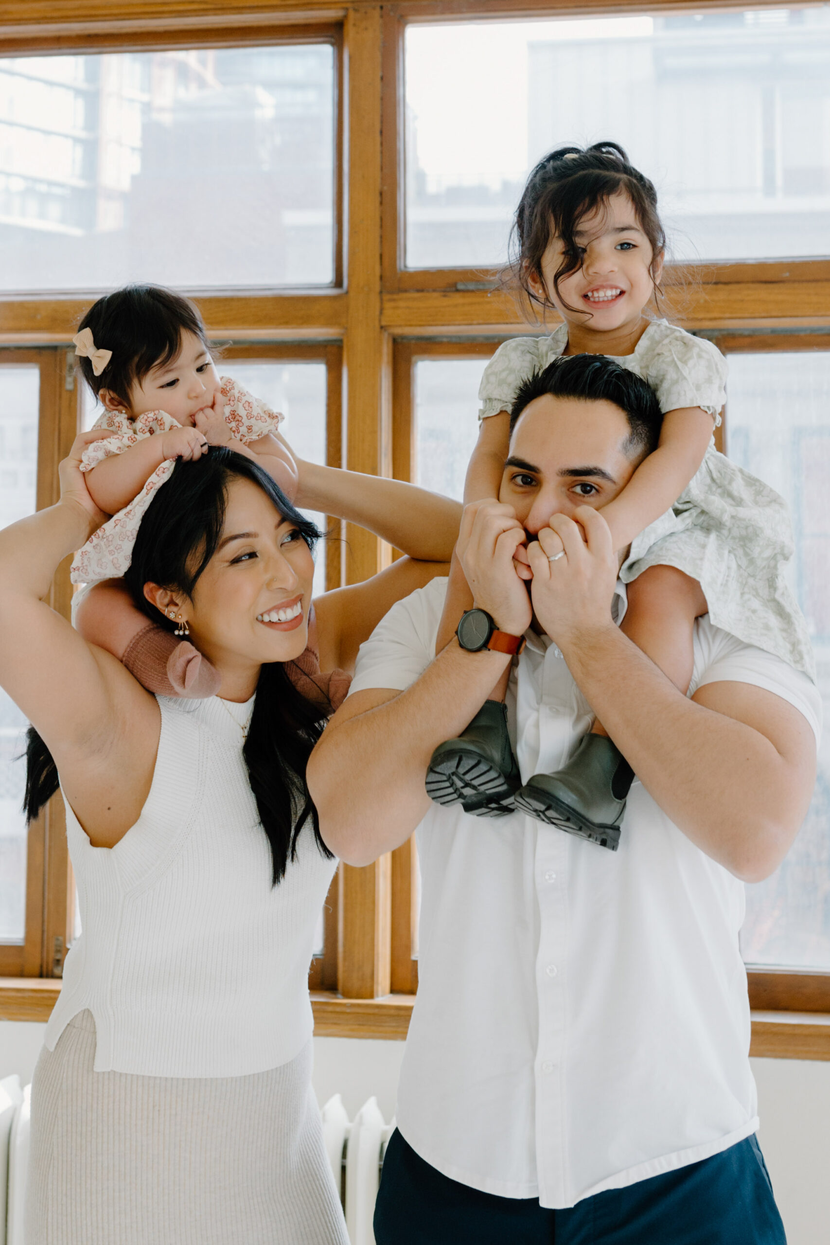 Two young girls sit on the mom and dad's shoulders by the windown