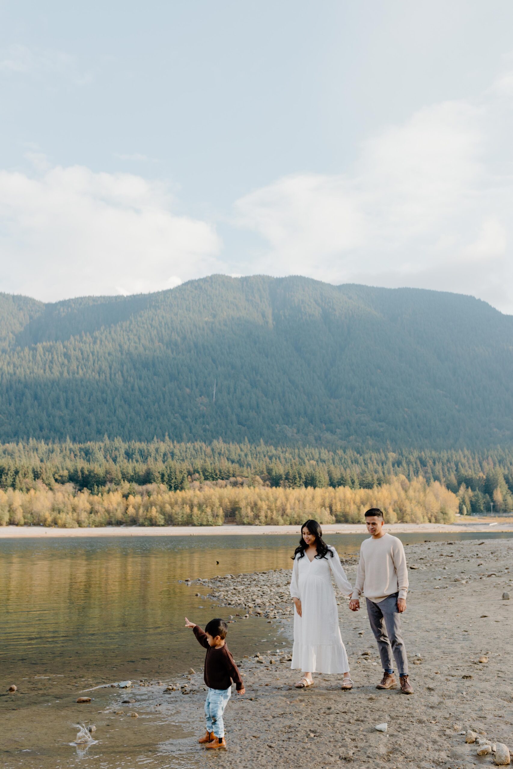 mom, dad and toddler boy are walking on the beach with mountains in the background